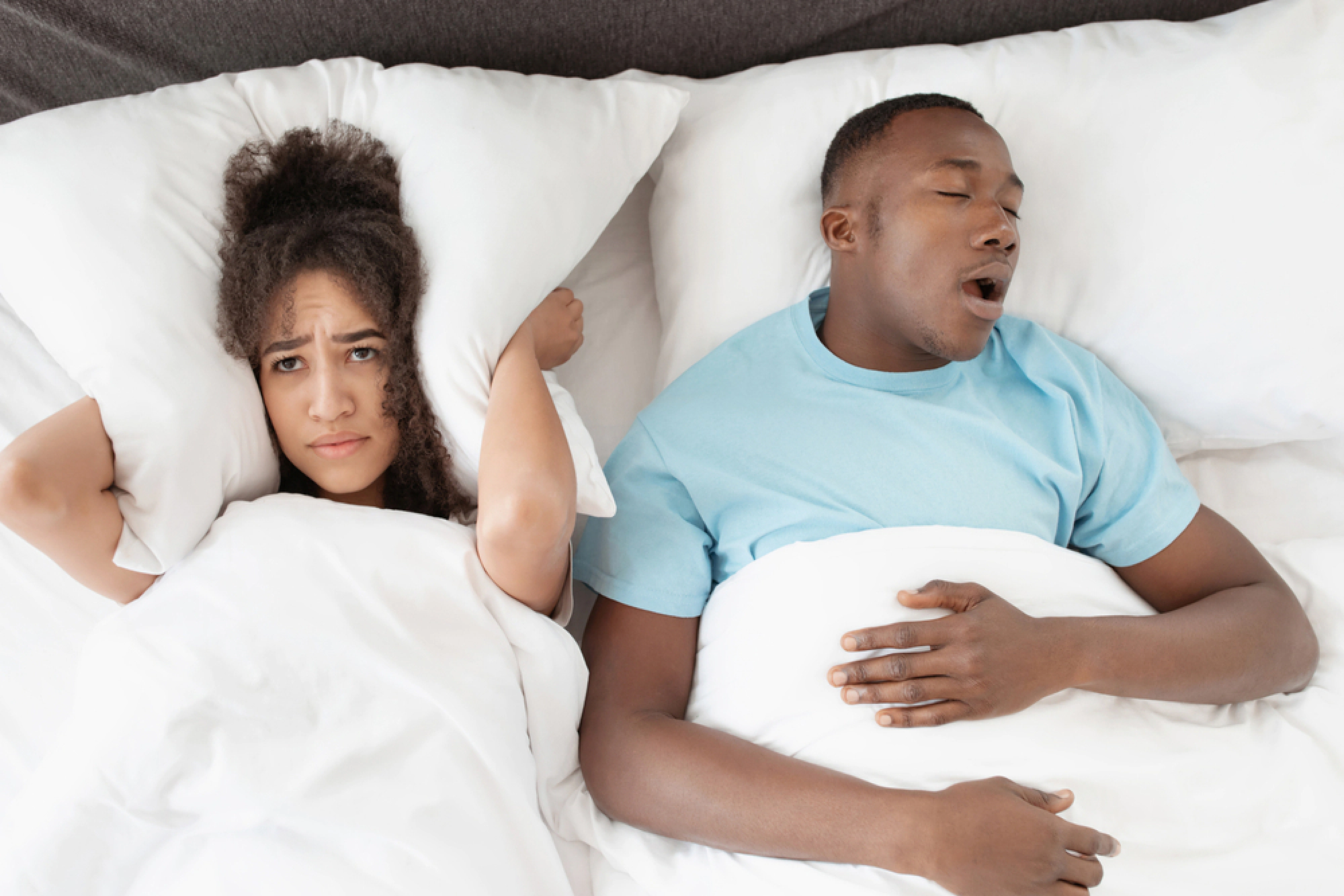 Husband snoring in bed while wife covers her ears with a pillow.