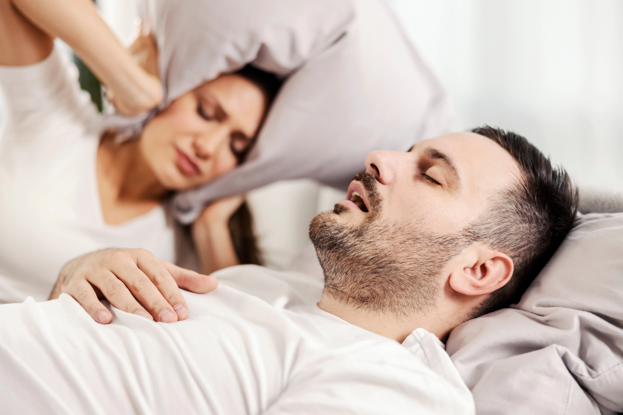 Man in bed snored while the woman next to him plugs her ears.
