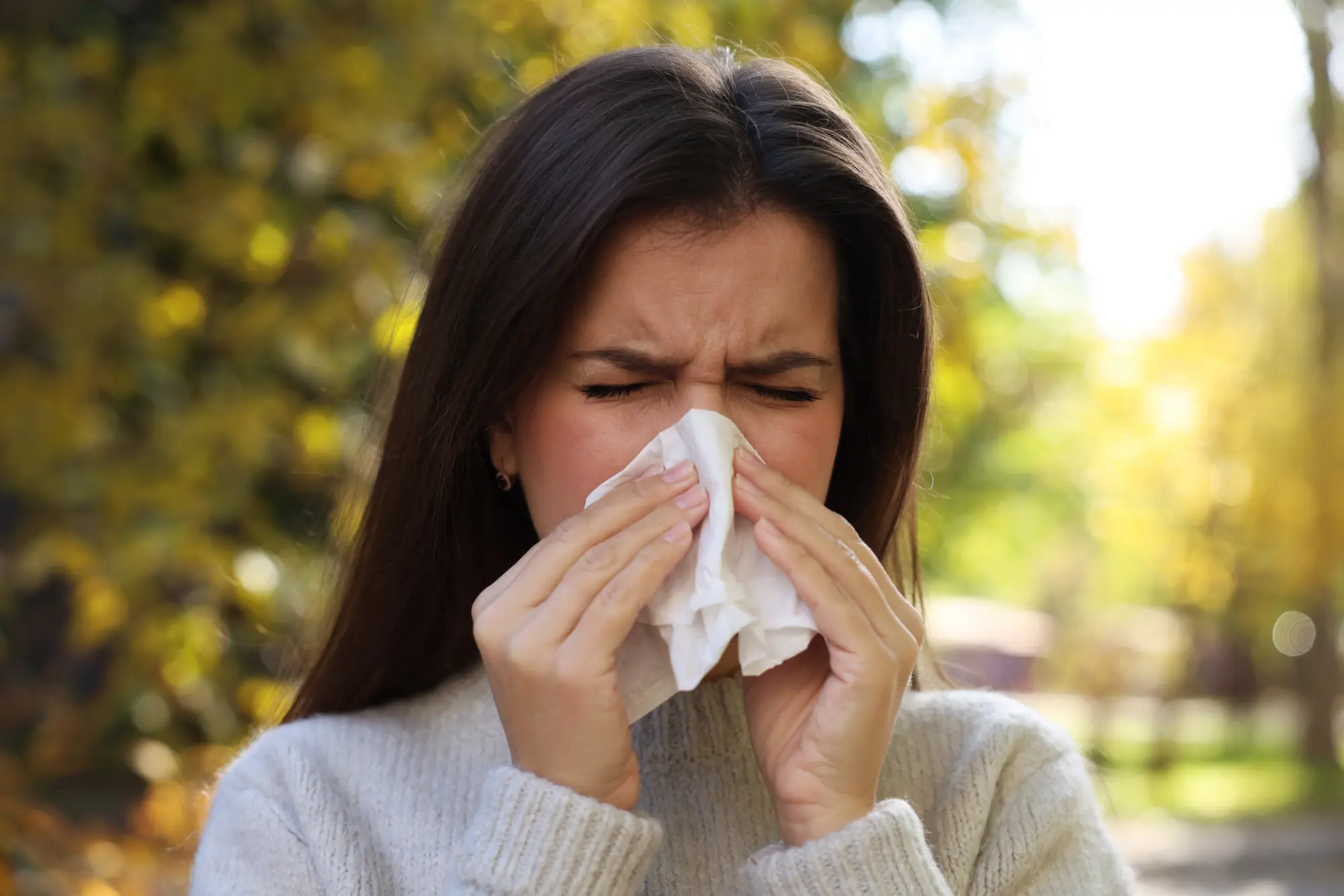 Woman sneezing into her tissue in front of fall trees